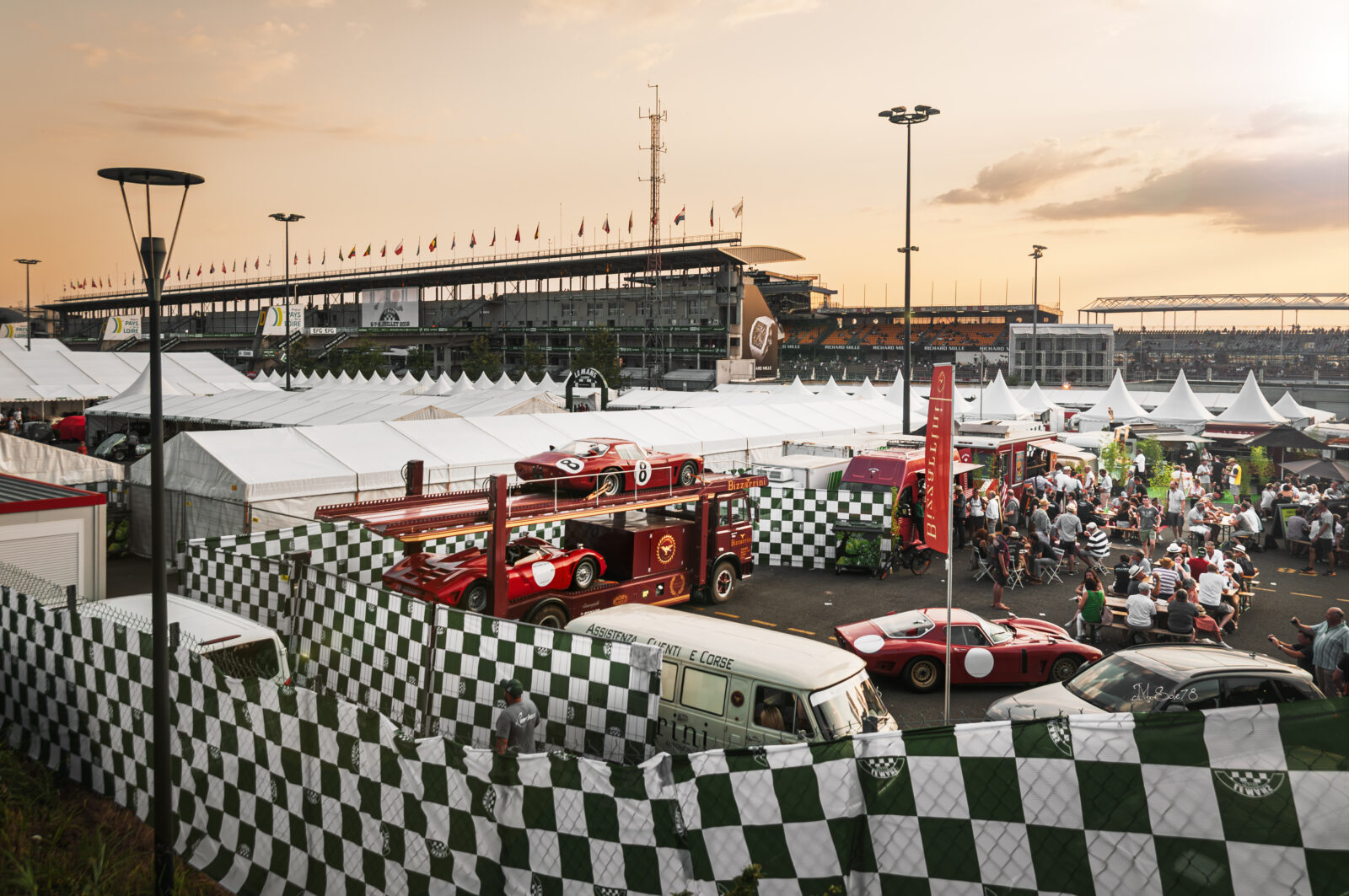 Auto da corsa rosse d'epoca nel paddock della 24 Ore di Le Mans al tramonto tra bandiere a scacchi